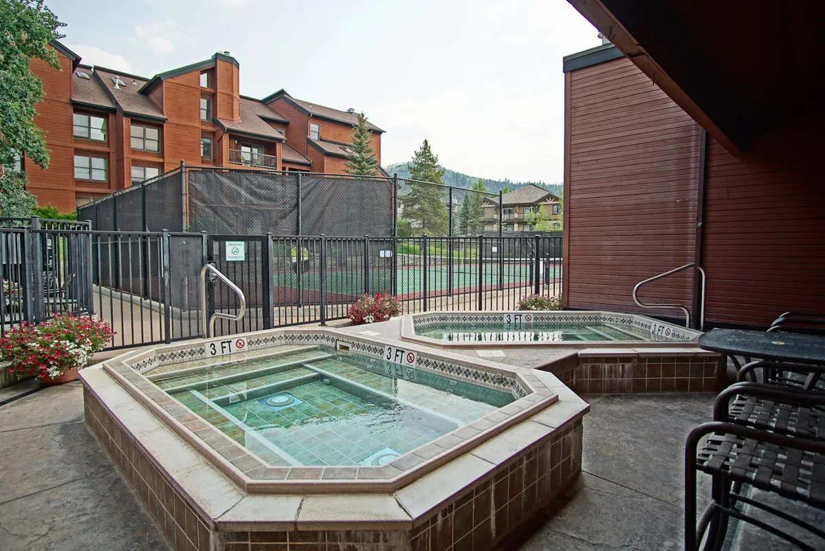 An exterior view of the hot tubs flanked by lush landscaping, with the fenced tennis court and part of the multi-story building visible in the background. The scene exudes relaxation and offers a glimpse of both water and sports-focused amenities.