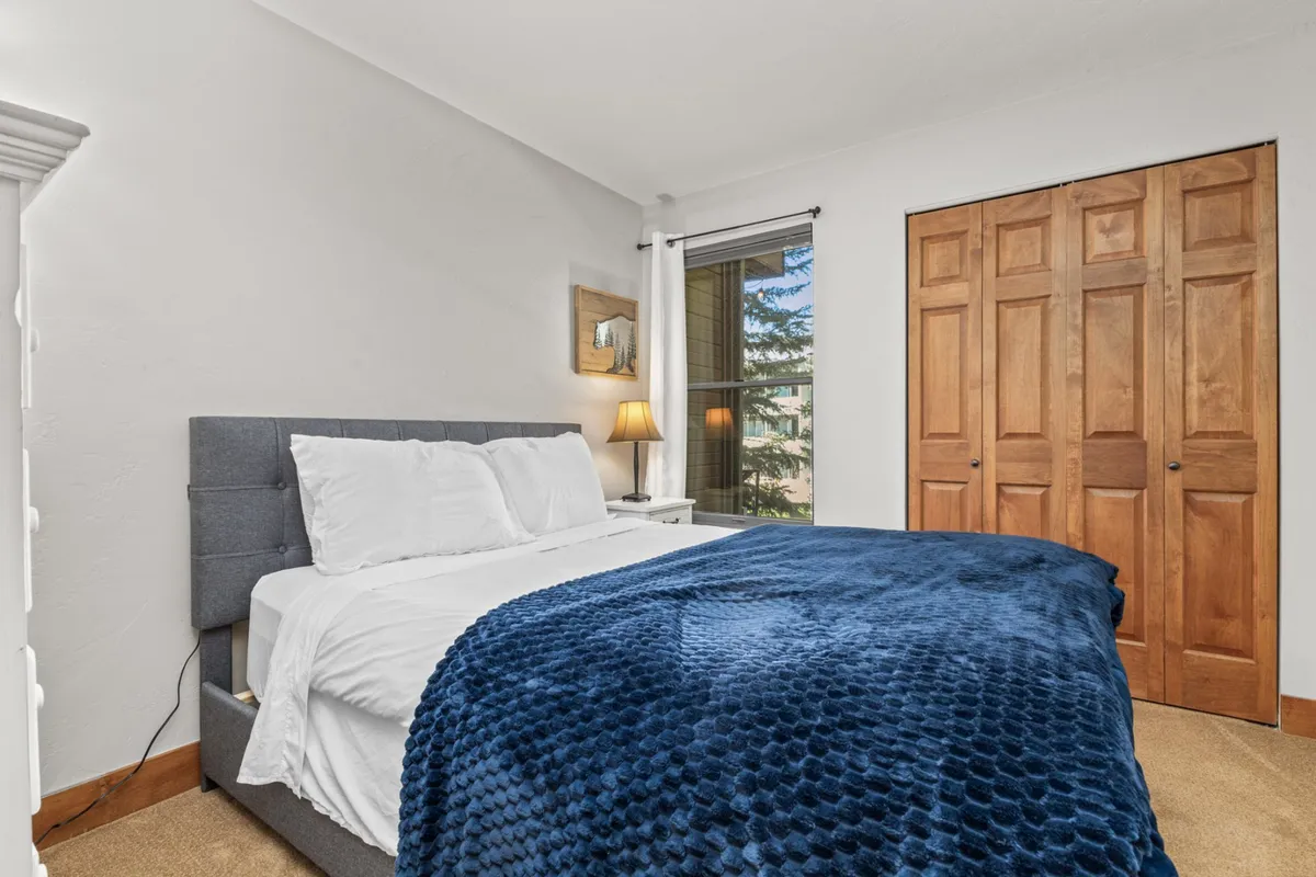 A bright and tranquil bedroom featuring a bed dressed with a blue throw blanket, natural light streaming through a window, wooden closet doors, and a softly glowing table lamp. The space feels airy and inviting—ideal for restful sleep and quiet moments.