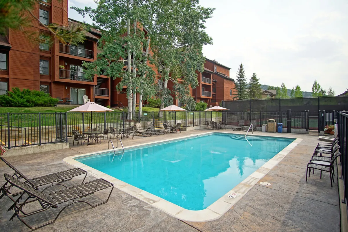 A vibrant outdoor swimming pool is surrounded by lush green trees, with several comfortable deck chairs and umbrellas lining the poolside. The area is inviting for sunbathing, swimming, and group relaxation, encapsulating the heart of the resort's summer amenities.