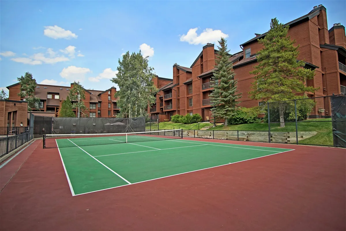 A well-maintained tennis court sits amidst a backdrop of leafy trees and adjacent resort buildings. The setting is peaceful but active, inviting guests to enjoy a friendly match or simply relax by the court.