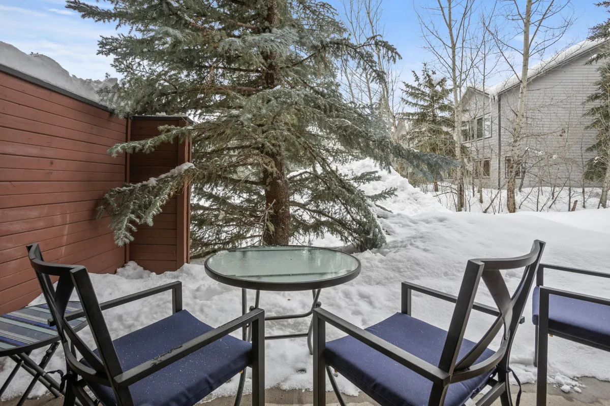 A snow-covered patio featuring a table and chairs nestled beneath a pine tree. The setting feels serene and wintry, inviting guests to enjoy the tranquil outdoors while surrounded by fresh powder after a day on the slopes.