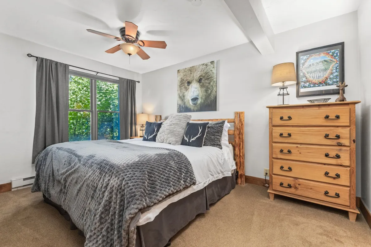 A cozy bedroom featuring a bed adorned with decorative pillows, charming bear artwork on the wall, a wooden dresser, and a window that frames lush greenery outside. The room is styled with soft, inviting linens and tasteful decor, creating a tranquil and comfortable ambiance perfect for unwinding.
