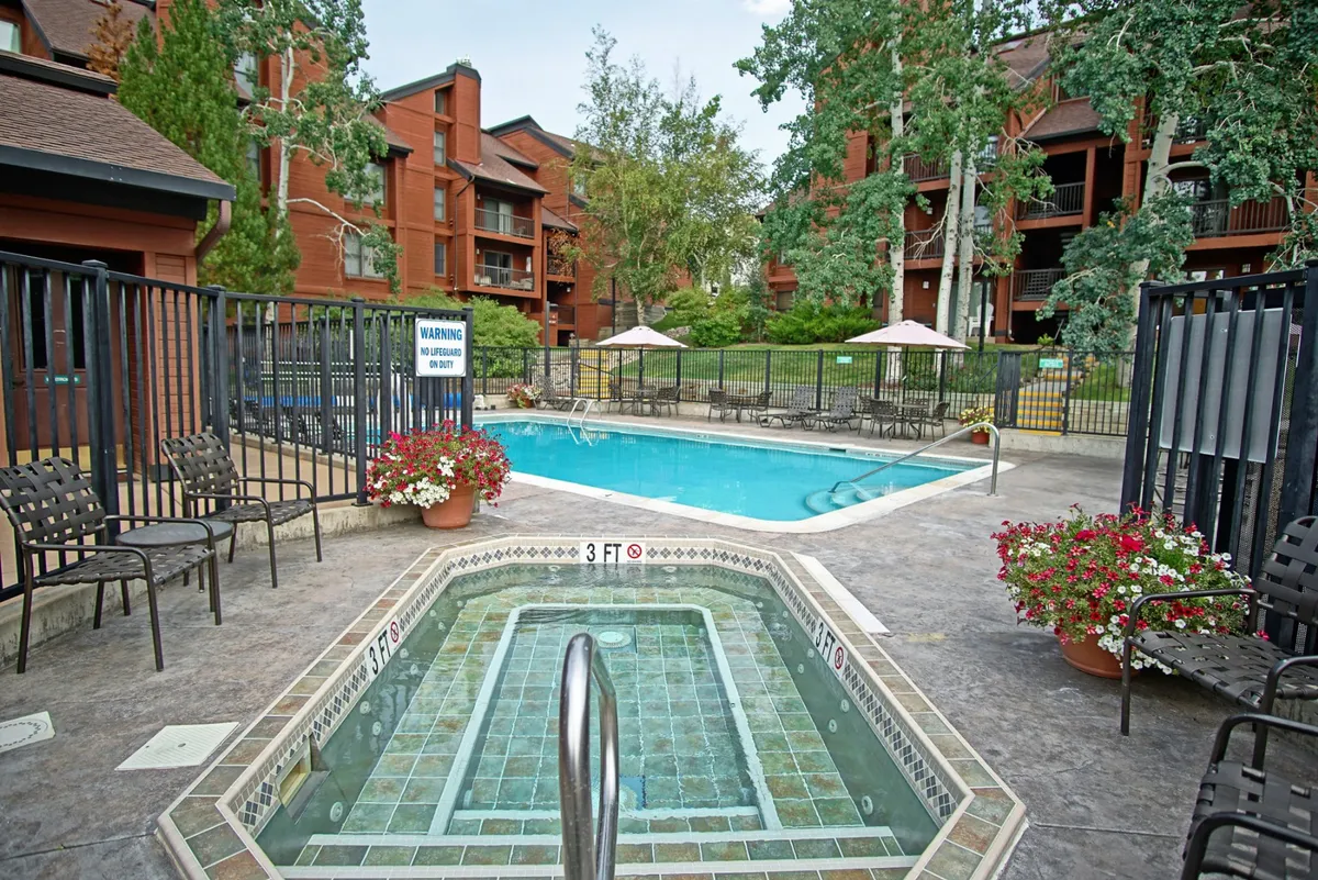 A vibrant exterior scene featuring both a sparkling heated pool and a bubbling hot tub, surrounded by lush greenery and comfortable lounge chairs. The setting is inviting and perfect for both relaxation and socializing under the open sky.