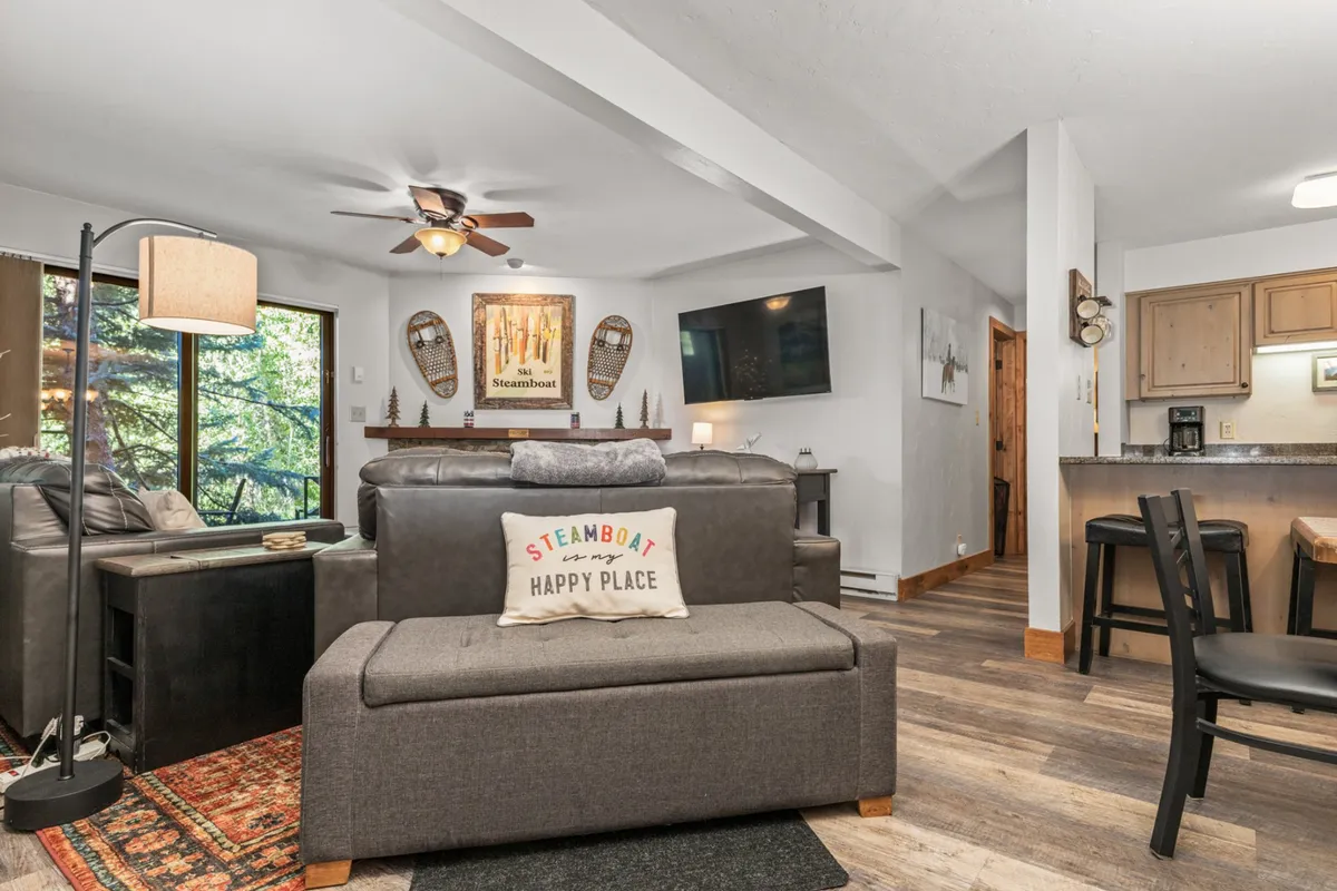 A cozy living room featuring a comfortable sofa with a 'Steamboat Happy Place' cushion, a wall-mounted television, decorative snowshoe wall art, and large windows filling the space with natural light. The arrangement invites relaxation and socializing, highlighting the open-concept's welcoming atmosphere.