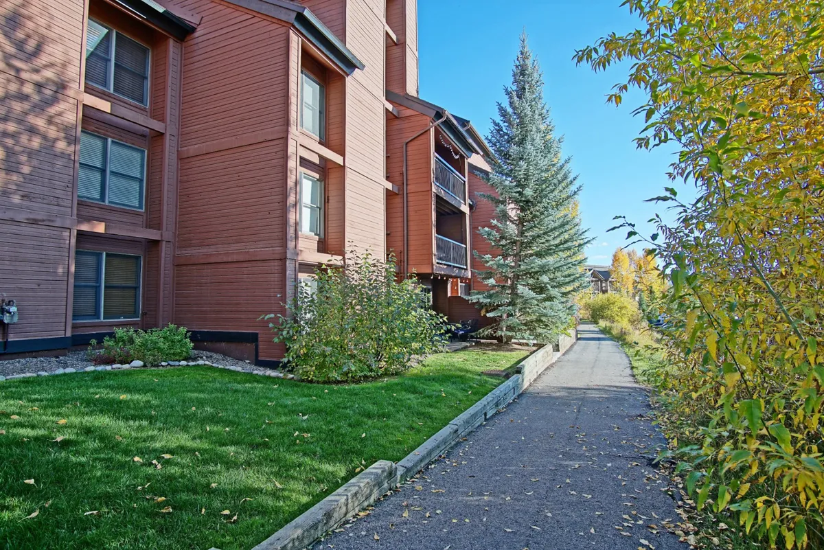 A tranquil exterior shot featuring the wooden siding of the building, a green lawn, a winding pathway, and vibrant autumn foliage. The setting is peaceful, inviting outdoor relaxation and easy walks to nearby destinations.