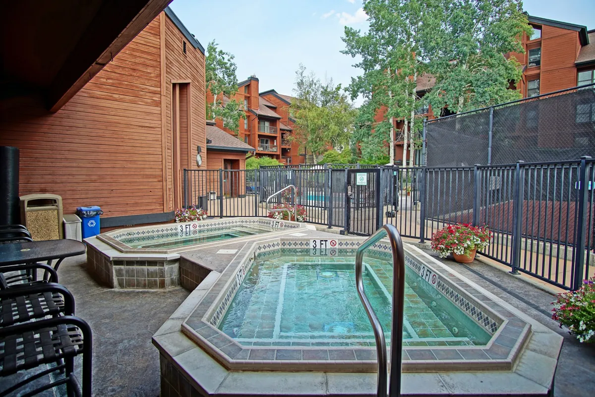 A fenced-in area with multiple inviting hot tubs set against lush greenery. The setting suggests a communal space to unwind and socialize, with the promise of warm relaxation after outdoor winter activities.