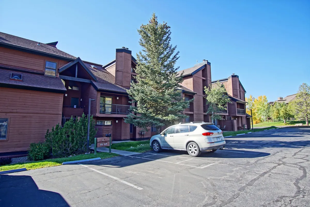 This exterior image shows the welcoming wooden exterior of the building, a tall pine tree, and a parked car, with balconies hinting at vibrant shared outdoor spaces. The landscaping and visible car emphasize accessibility and the possibility of socializing outdoors.